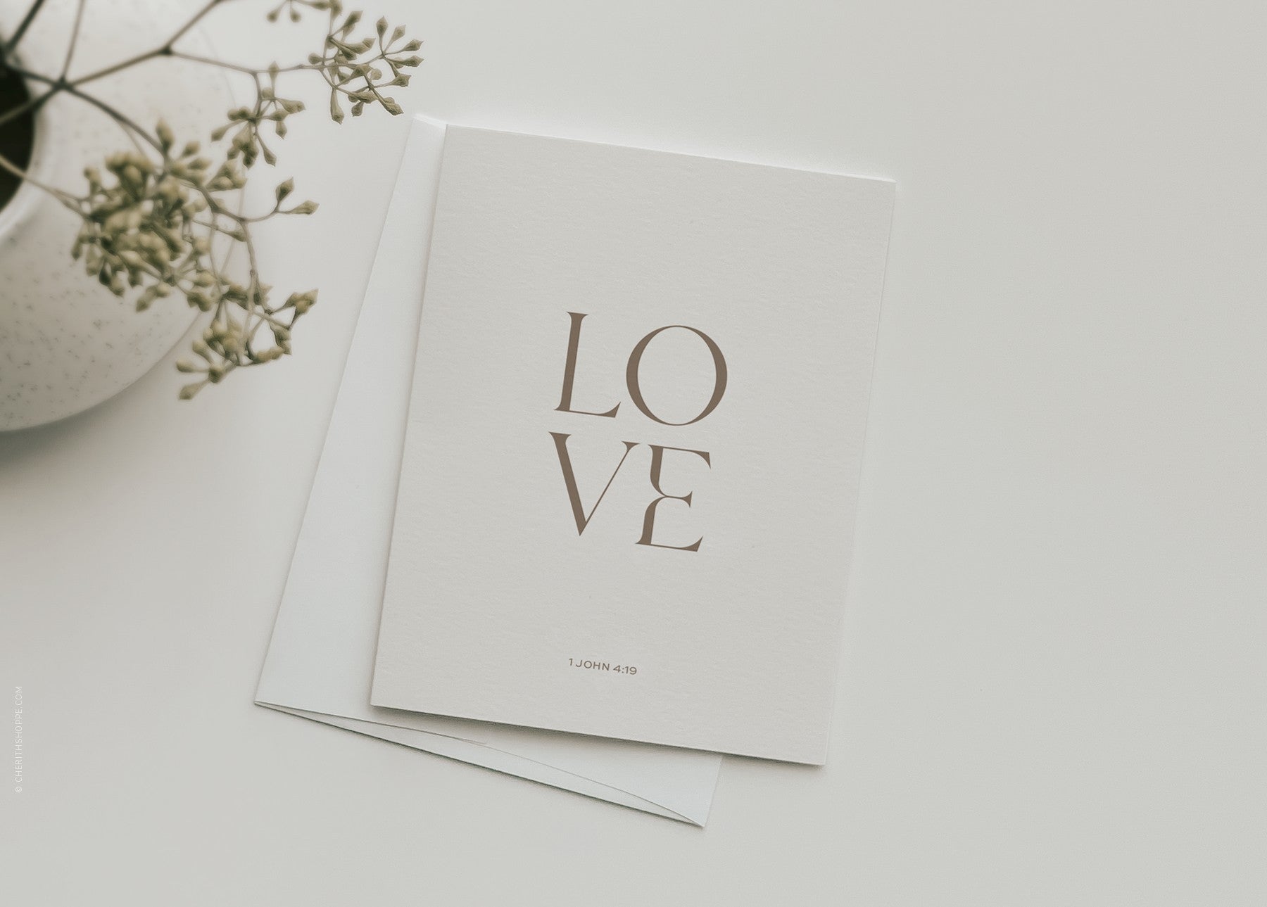 “Love” 1 John 4:19 Christian greeting card by Cherith with a white envelope, styled on a white table.
